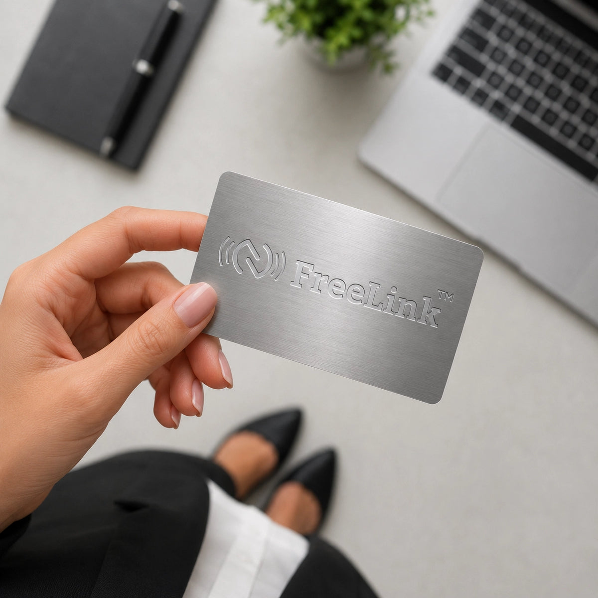 Hand holding a metallic silver NFC metal card with 'Freelink' branding on a desk with laptop and pen.